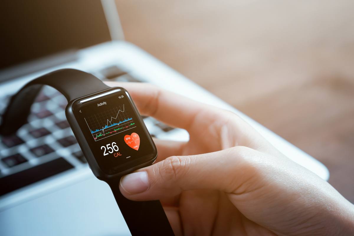 Close-up of a smartwatch on a person's wrist displaying heart rate and calories burned, with a blurred laptop in the background.