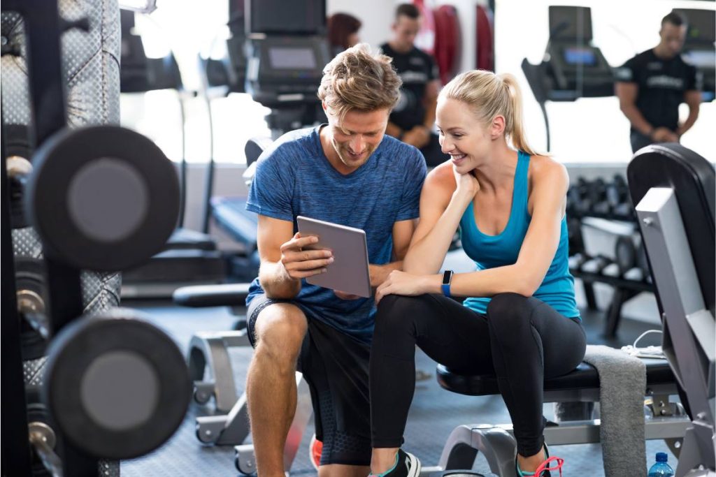 Two people looking at a tablet in a gym, with workout equipment in the background.